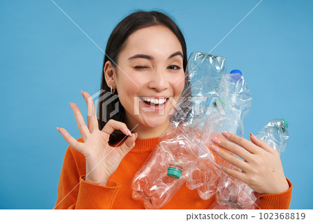 Recycling is great. Smiling young woman, holding bottles, shows okay sign, approves eco green lifestyle, sorting garbage, blue background Recycling is great. Smiling young woman, holding bottles, shows okay sign, approves eco green lifestyle, sorting garbage, blue background 102368819
