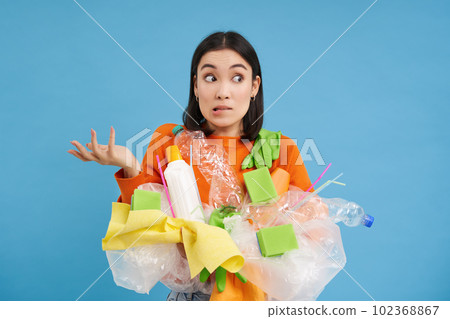 Korean girl with plastic garbage, shrugs shoulders, doesnt know how to recycle, sorting her waste for recycling station, blue background 102368867