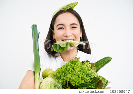Portrait of smiling korean woman eats lettuce leaf, holding green vegetables, eating nutritious food, white background Portrait of smiling korean woman eats lettuce leaf, holding green vegetables, eating nutritious food, white background 102368947