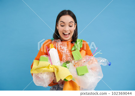 Happy girl looks at plastic garbage, sorting her waste for recycling station, smiling excited, blue background. Environment and ecology concept 102369044