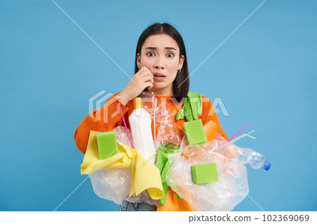 Woman looks scared and anxious, holds plastic waste, nervous to recycle properly, blue studio background Woman looks scared and anxious, holds plastic waste, nervous to recycle properly, blue studio background 102369069