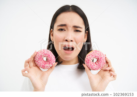 Close up portrait of sad asian woman, upset being on diet, showing two glazed pink doughnuts, tempted to eat junk food, white background Close up portrait of sad asian woman, upset being on diet, showing two glazed pink doughnuts, tempted to eat junk food, white background 102369153