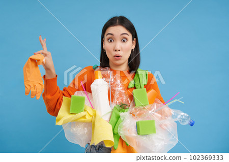 Asian woman looks shocked, holds plastic rubbish, sorting garbage for recycling, looks surprised, blue background 102369333
