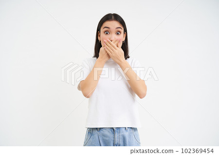 Portrait of girl with shocked face, holds hands on her mouth, stares speechless, startled by gossip, white background 102369454