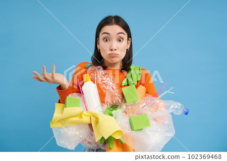 Korean girl with plastic garbage, shrugs shoulders, doesnt know how to recycle, sorting her waste for recycling station, blue background Korean girl with plastic garbage, shrugs shoulders, doesnt know how to recycle, sorting her waste for recycling station, blue background 102369468