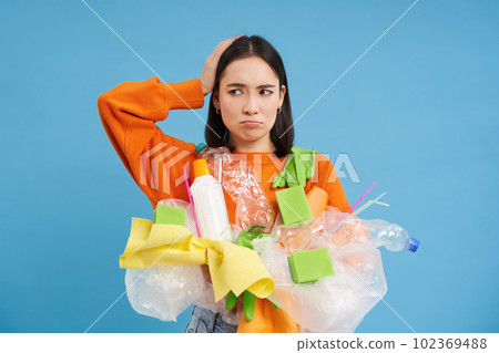 Confused girl with plastic garbage in hands, scratches head, looks puzzled with recycling system, blue background 102369488