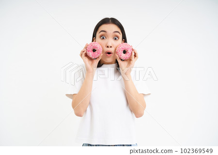 Beautiful young woman eating delicious glazed dougnuts, holding two pink donuts and smiling, white background Beautiful young woman eating delicious glazed dougnuts, holding two pink donuts and smiling, white background 102369549
