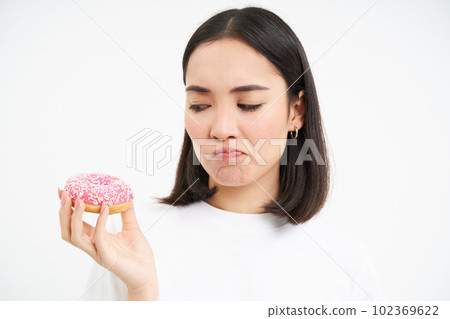 Close up of sad cute girl, shows tasty glazed doughnut, being on diet, cant try delicious donnut, junk food concept Close up of sad cute girl, shows tasty glazed doughnut, being on diet, cant try delicious donnut, junk food concept 102369622