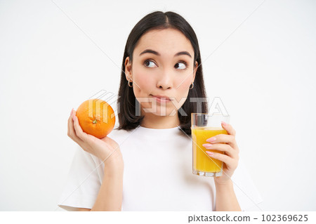 Close up portrait of asian woman with orange and glass of juice, thinking, making choice, standing over white background 102369625
