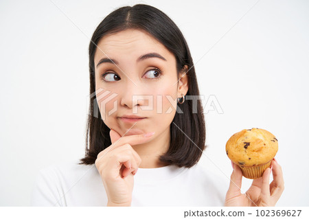Close up portrait of korean woman with one cupcake, thinking and smiling, white studio background 102369627