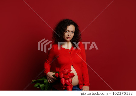 Multi ethnic curly haired pregnant woman looking confidently at camera, holding a bouquet of red tulips on isolated color background, dressed in bright red shirt and blue jeans. Pregnancy fashion 102370152