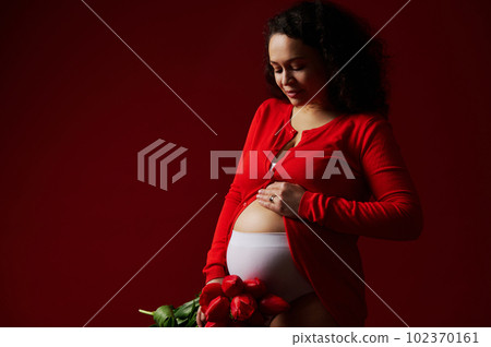 Fashion portrait shot in low key of delightful happy pregnant woman, expectant mother caressing her belly, smiling posing with a bouquet of red tulips on isolated red background. Pregnancy. Maternity 102370161