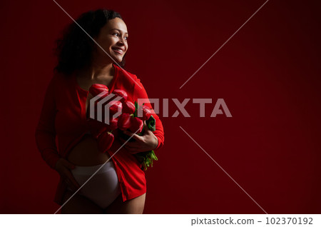 Happy adult pregnant woman caressing her belly, smiling broadly with a beautiful toothy smile, looking aside, posing with a bouquet of tulips over isolated red color background. Copy space. Pregnancy 102370192