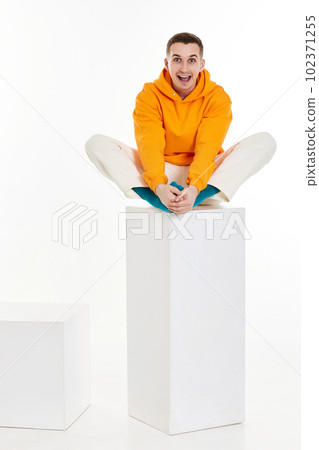 young man in orange sweatshirt sitting on white cube young man in orange sweatshirt sitting on white cube 102371255