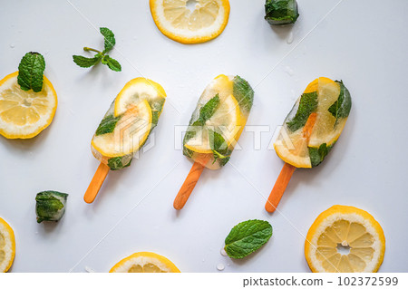 three appetizing pieces of fruity lemon mint ice on sticks lie on a light background with citrus slices and mint leaves 102372599