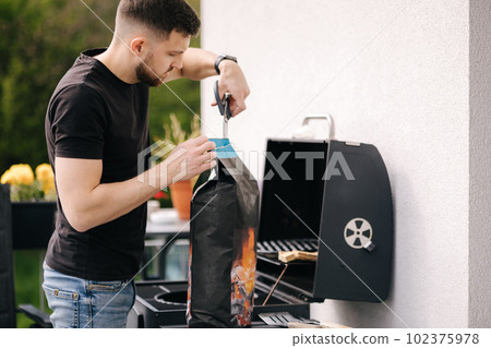 Man opens a package of charcoal for grilling. Preparing for barbecue on backyard  102375978