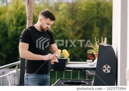 Portrait of handsome young man stand by BBQ grill on terrace and wipes hand  102375994