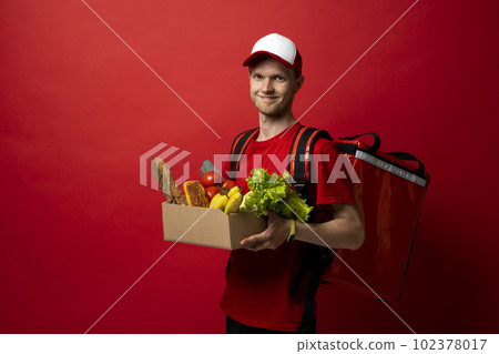 Delivery male employee in a red uniform holds paper cardboard package with organic groceries, fruits on a red background. Products delivery from shop or restaurant to home. Courier service. 102378017