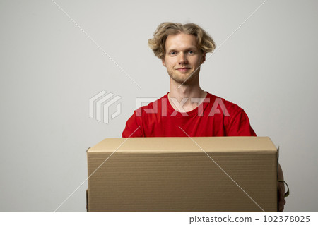 Delivery service. Young smiling courier holding cardboard box. Happy young delivery man in red t-shirt standing with parcel isolated on white background. 102378025