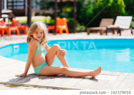 Cute and funny blonde teenage girl in a swimsuit sits on the side of the pool.  102379058