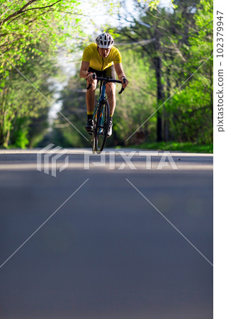 Young man in protective helmet and activewear biking on paved road in a green forest. Vertical photo. Young man in protective helmet and activewear biking on paved road in a green forest. Vertical photo. 102379947