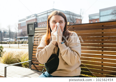 Unhappy caucasian millennial red-haired woman sneezing in napkin and siting in park bench, Young women suffering from allergy outdoors Unhappy caucasian millennial red-haired woman sneezing in napkin and siting in park bench, Young women suffering from allergy outdoors 102382454