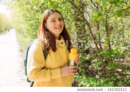 Happy redhead Woman drinking water from water bottle or hot drink from yellow thermos on nature background. Smile pretty girl with freedom time Happy redhead Woman drinking water from water bottle or hot drink from yellow thermos on nature background. Smile pretty girl with freedom time 102382812