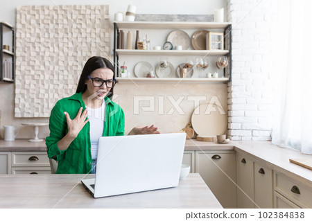 Portrait of an upset woman sitting at home in the kitchen and talking unhappily through a video call. Arguing, discussing, indignant. 102384838