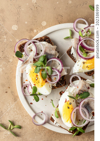 Smorrebrod sandwiches with herring fillet, rye bread, cream cheese, boiled egg slices, onion rings and microgreens on white dish brown background, rustic style. Top view Smorrebrod sandwiches with herring fillet, rye bread, cream cheese, boiled egg slices, onion rings and microgreens on white dish brown background, rustic style. Top view 102386999