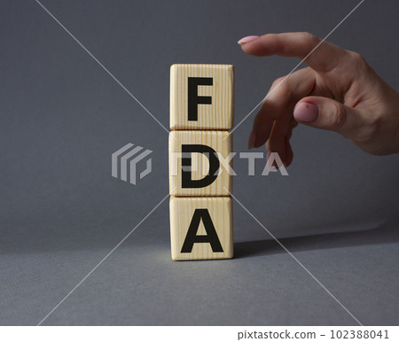 FDA - Food Drug Administration symbol. Wooden cubes with word FDA. Doctor hand. Beautiful grey background. Medical and Food Drug Administration concept. Copy space. FDA - Food Drug Administration symbol. Wooden cubes with word FDA. Doctor hand. Beautiful grey background. Medical and Food Drug Administration concept. Copy space. 102388041