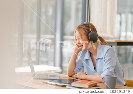 Happy Asian businesswoman wearing headphones is taking notes in a notebook while watching a webinar video course. happy female student listening to the lecture to study online through e-learning. Happy Asian businesswoman wearing headphones is taking notes in a notebook while watching a webinar video course. happy female student listening to the lecture to study online through e-learning. 102391249