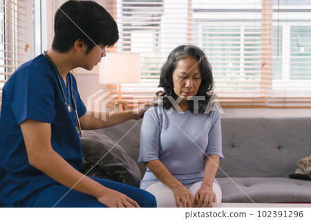 A young caregiver is consulting with a 60-year-old Asian elderly woman, providing encouragement and taking a medical history. The two are shown holding hands and exchanging words of encouragement. 102391296