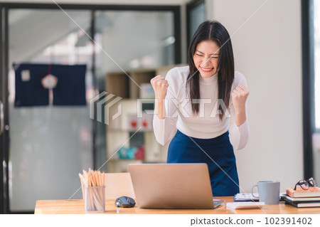 Happy and excited, a young asian woman student or office worker, celebrates achieving her goal of winning an online competition or receiving good news via email by raising hands in euphoria. 102391402