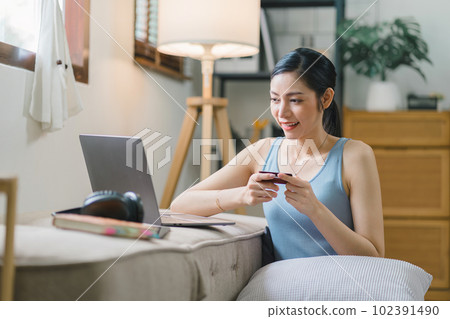 A happy young woman is shown shopping online using her laptop and credit card at home. 102391490