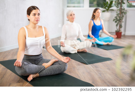 Calm women of different ages meditating in Lotus Pose or Padmasana in beginning yoga training together in cozy gym studio Calm women of different ages meditating in Lotus Pose or Padmasana in beginning yoga training together in cozy gym studio 102392295