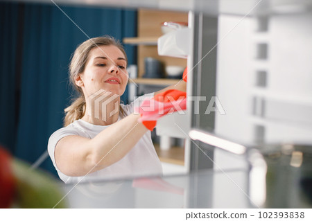 Portrait of a woman cleaning in a kitchen and wearing red gloves. Caucasian woman doing housework and standing in front of opened refrigerator. View from inside of kitchen refrigerator. 102393838