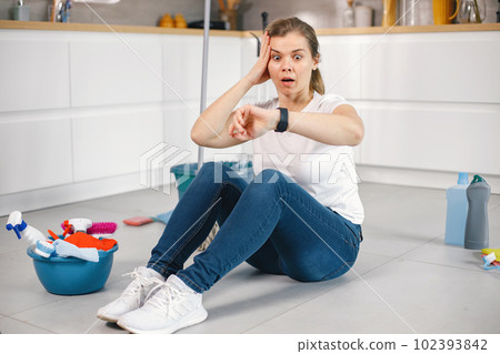 Portrait of a woman sitting in a kitchen on a floor and wearing red gloves. Caucasian woman after doing housework. Adult woman looking on her watch and have surprised face. Portrait of a woman sitting in a kitchen on a floor and wearing red gloves. Caucasian woman after doing housework. Adult woman looking on her watch and have surprised face. 102393842