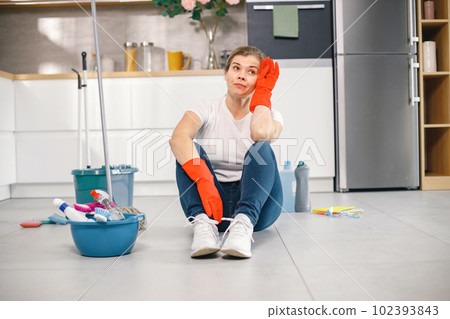 Portrait of a woman sitting in a kitchen on a floor and wearing red gloves. Caucasian woman after doing housework. Adult woman looking tired. 102393843