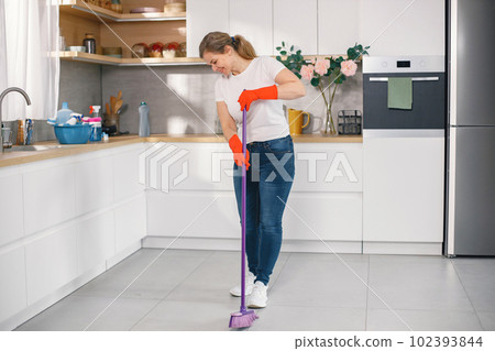 Portrait of a woman cleaning in a kitchen and wearing red gloves. Caucasian woman hdoing housework. Adult woman sweeping a floor with a broom. 102393844