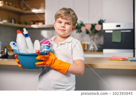 Portrait of a blonde boy cleaning in a kitchen and wearing orange gloves. Caucasian boy helping his mother to do housework. Boy holding blue busket with different rugs and detergents. Portrait of a blonde boy cleaning in a kitchen and wearing orange gloves. Caucasian boy helping his mother to do housework. Boy holding blue busket with different rugs and detergents. 102393845