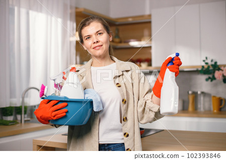Portrait of a woman cleaning in a kitchen and wearing red gloves. Caucasian woman doing housework. Adult woman holding blue busket with different rugs and detergents. Portrait of a woman cleaning in a kitchen and wearing red gloves. Caucasian woman doing housework. Adult woman holding blue busket with different rugs and detergents. 102393846