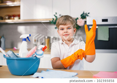 Blonde boy cleaning in a kitchen and wearing orange gloves. Caucasian boy helping his mother to do housework. Blue busket with different rugs and detergents on a table. Blonde boy cleaning in a kitchen and wearing orange gloves. Caucasian boy helping his mother to do housework. Blue busket with different rugs and detergents on a table. 102393858