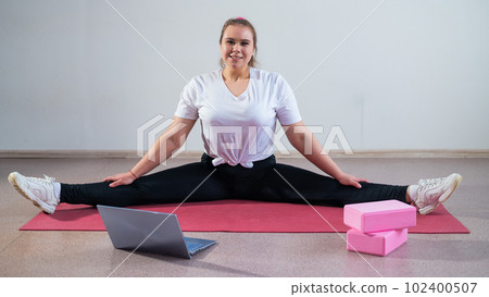 Young caucasian fat woman doing bends on a sports mat and watching a training video on a laptop. A chubby girl stretches the split remotely using video communication Young caucasian fat woman doing bends on a sports mat and watching a training video on a laptop. A chubby girl stretches the split remotely using video communication 102400507