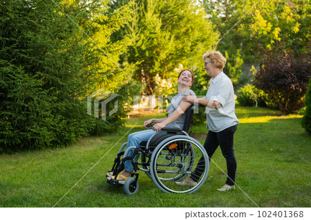 An elderly woman is carrying an adult daughter sitting in a wheelchair. Walk outdoors.  102401368