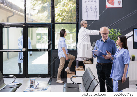 Medical assistant explaining disease expertise to elderly patient while discussing health care treatment during checkup visit consultation in hospital lobby. Medicine service and concept 102403783
