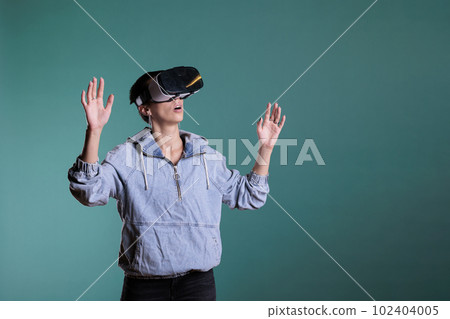 Cheerful fun woman wearing virtual reality goggles with video interaction, standing over blue background. Confident adult playing videogames using vr technology with 3d tech simluation 102404005