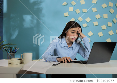 Asian woman sitting at office desk suffering from severe headache while working with laptop computer. Exhausted female corporate employee in pain looking tired from so much work. 102404077