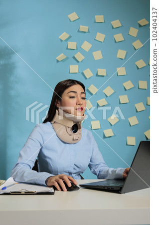 Asian woman with cervical collar on neck using laptop computer at modern office desk. Female employee with injury making an effort to finish sales reports in startup company. Asian woman with cervical collar on neck using laptop computer at modern office desk. Female employee with injury making an effort to finish sales reports in startup company. 102404137