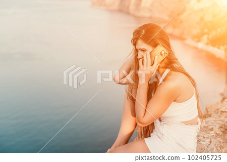 Happy woman in white shorts and T-shirt, with long hair, talking on the phone while enjoying the scenic view of the sea in the background. 102405725