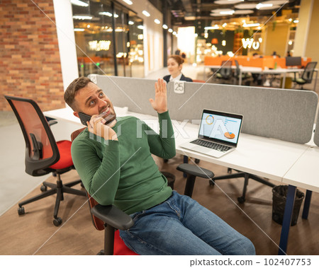 Caucasian bearded man talking on a smartphone while sitting at his desk in a modern coworking space. Caucasian bearded man talking on a smartphone while sitting at his desk in a modern coworking space. 102407753
